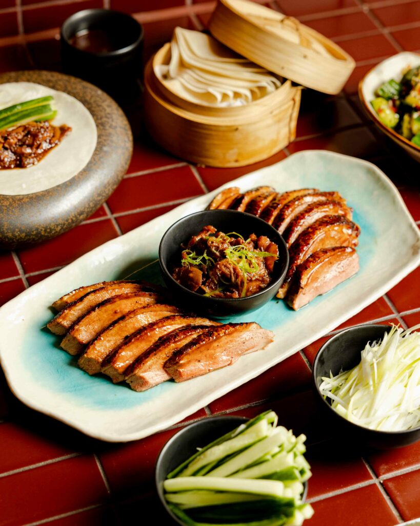 An elegant presentation of Peking duck, sliced and served on a blue ceramic plate, accompanied by steamed buns, sauces, and fresh green vegetables, arranged on a dark red tiled table.