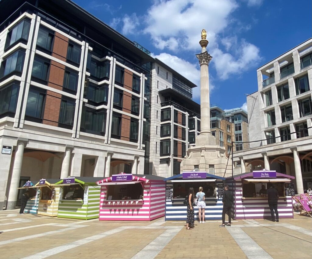 Paternoster Square - a large public space with a column in the middle, and surrounded by buildings. In this image there are brightly coloured food stalls from a summer market.