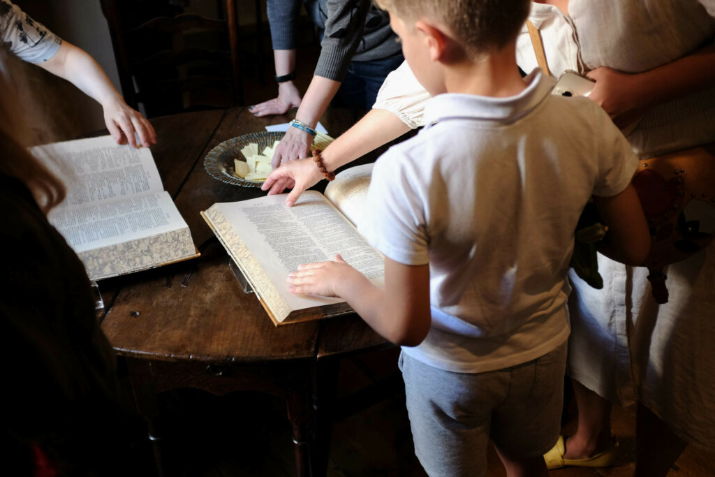 A group of people gathered around a wooden table, looking at an open large book. There is a plate with small scraps of paper on the table.