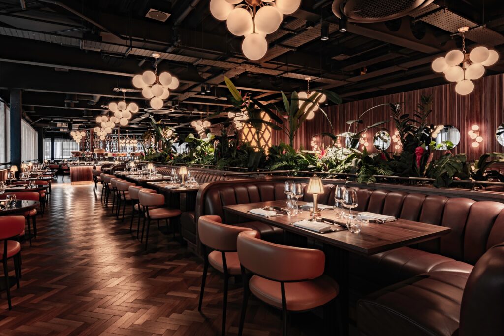 Interior of Fazenda Rodizio Bar & Grill at Bishopsgate featuring a stylish dining area with leather seating, wooden tables, and an expansive bar. Décor includes large, pendant lights and lush green plants.