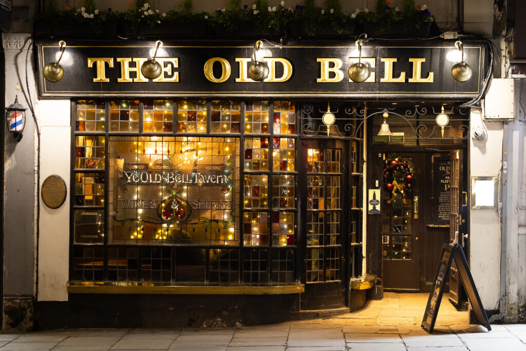 Exterior of the black painted Old Bell Tavern taken at night. The large windows are lit up by a light inside.