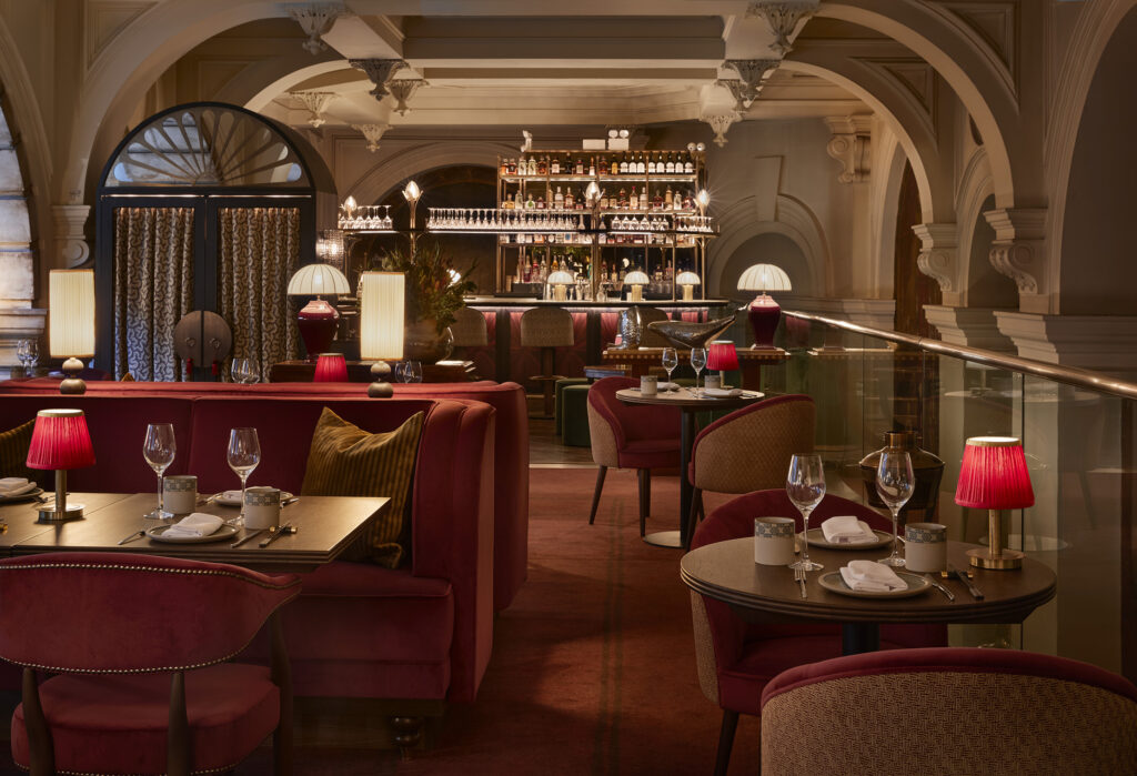 Elegant interior of a dimly lit restaurant featuring plush red seating, white tablecloths on neatly set tables, and a grand, well-stocked bar at the back beneath arched white architectural details.