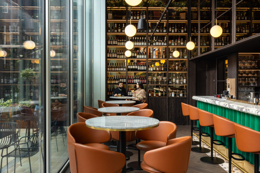 The interior of Dave's Wine Bar features a row of circular marble tables, velvet seating, and a floor-to-ceiling wooden bookshelf.