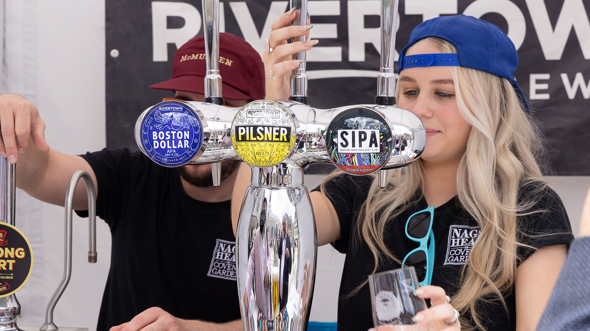Two people, one wearing a red cap with writing 'McMullen' and one wearing a black cap backwards pulling pints. The beer taps have the colourful logos of the beers with text overlayed 'Rivertown Boston Dollar', 'Rivertown Pilsner' 'SIPA Rivertown'.
