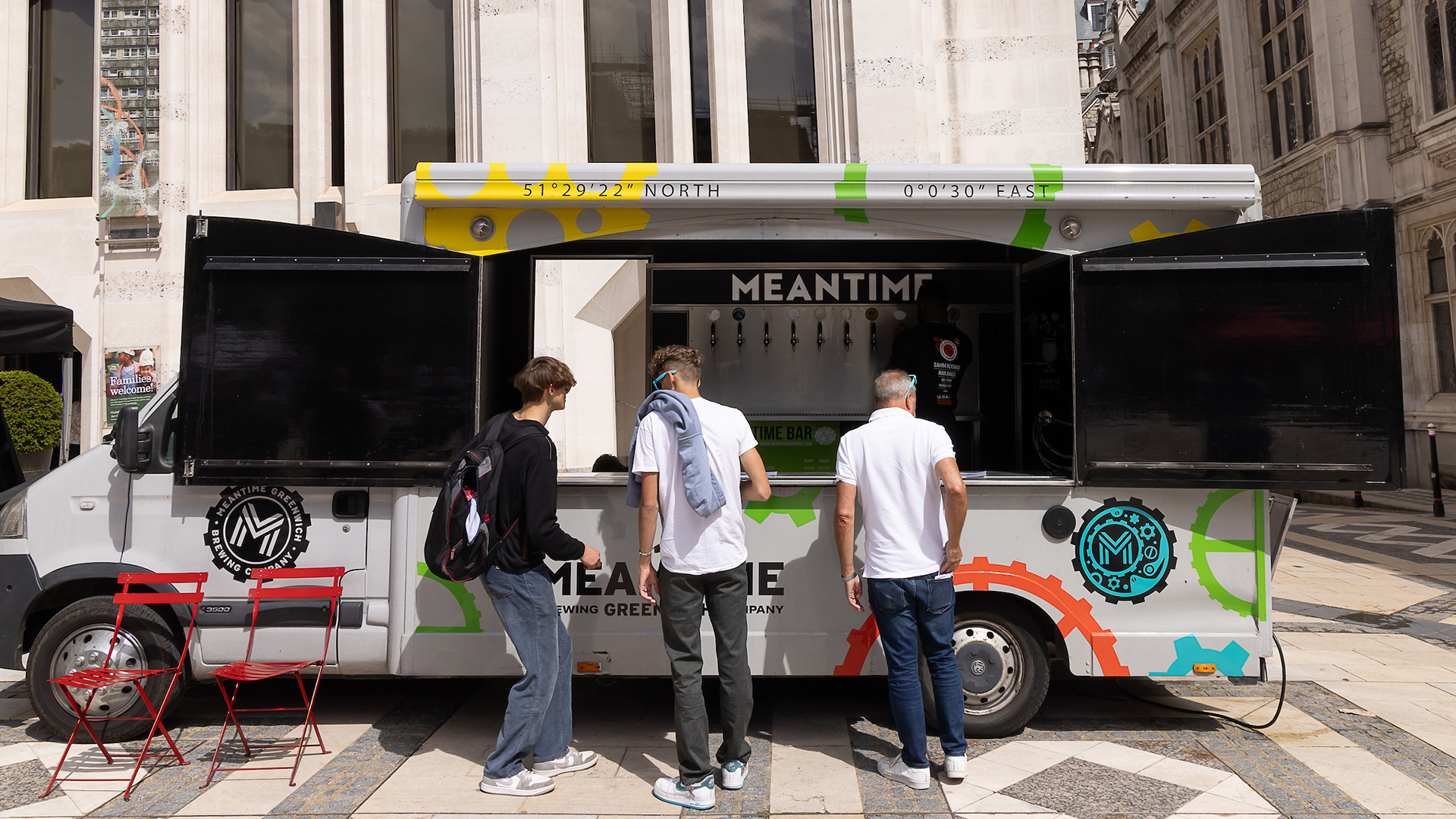 The Meantime Brewery Truck is white with a colourful wheels and cog design. The windows to the truck are open with the beer taps on display, a person pulling a pint, and three people waiting outside the truck.
