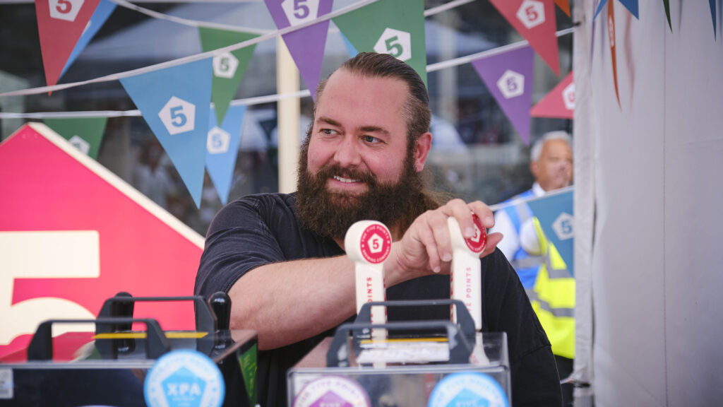 Person in black t-shirt and smiling is pulling a pint with colourful bunting in the background.