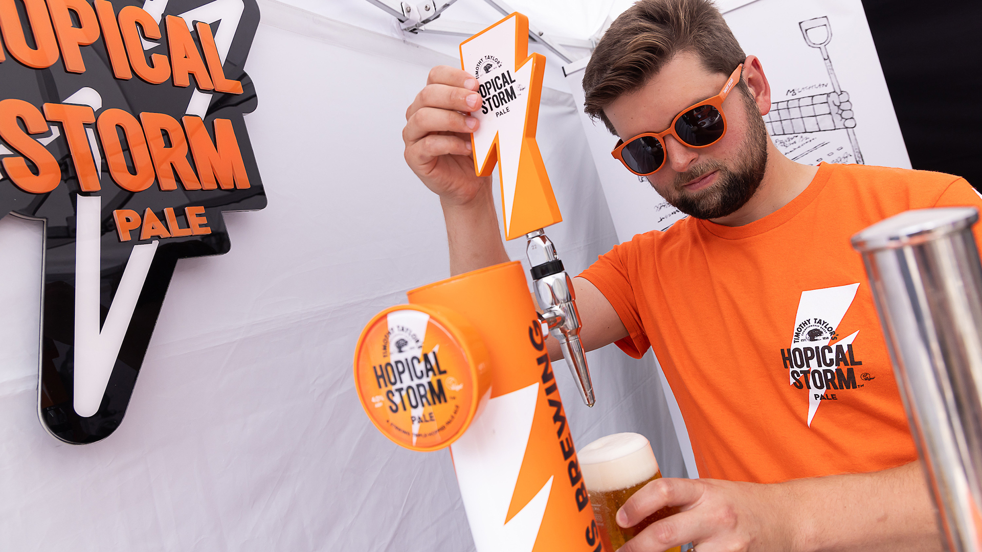 Person in orange t-shirt with a lightning logo and the words 'Hopical Storm Pale' overlayed. Person is pulling a pint in white tent.