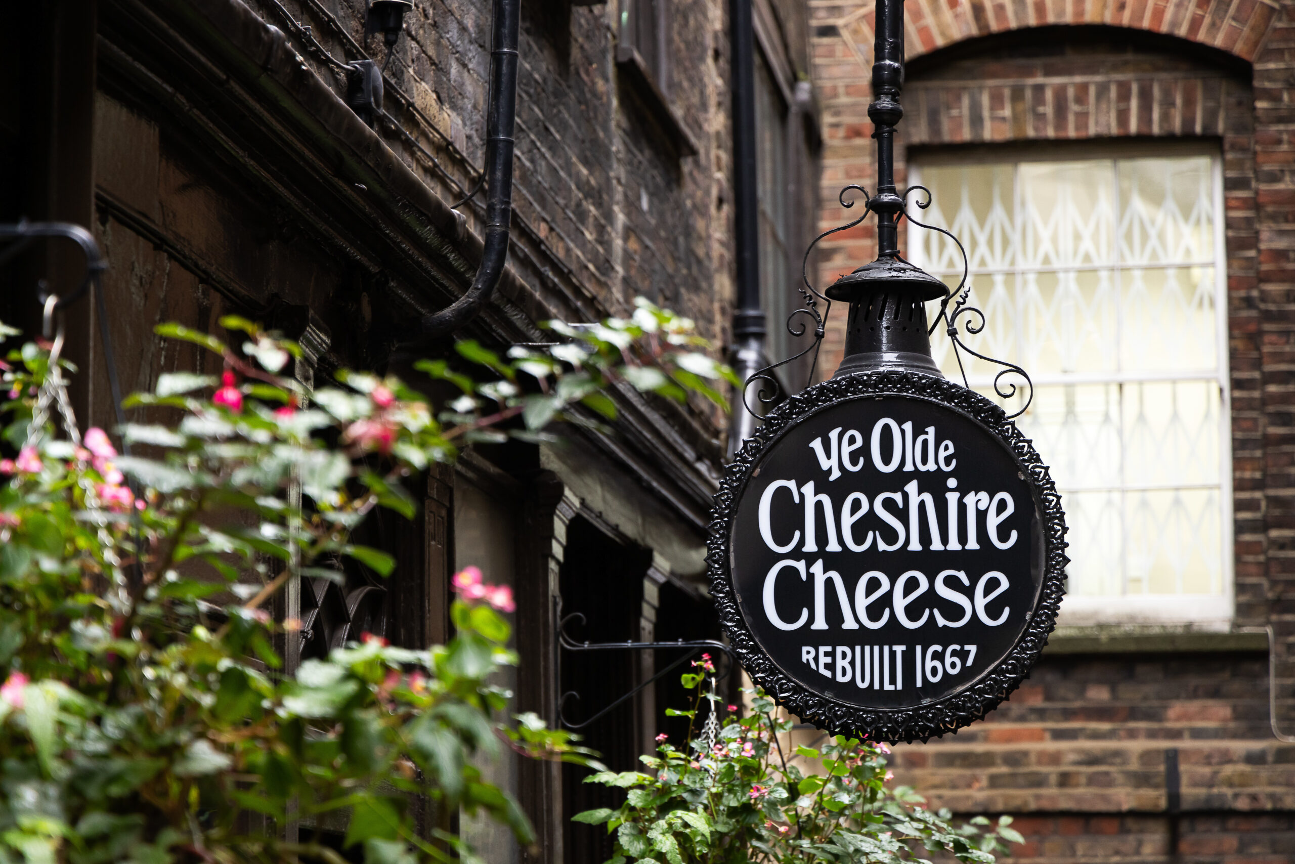 A black, round, hanging sign reading 'Ye Olde Cheshire Cheese' set against a backdrop of a red brick building and lush greenery.