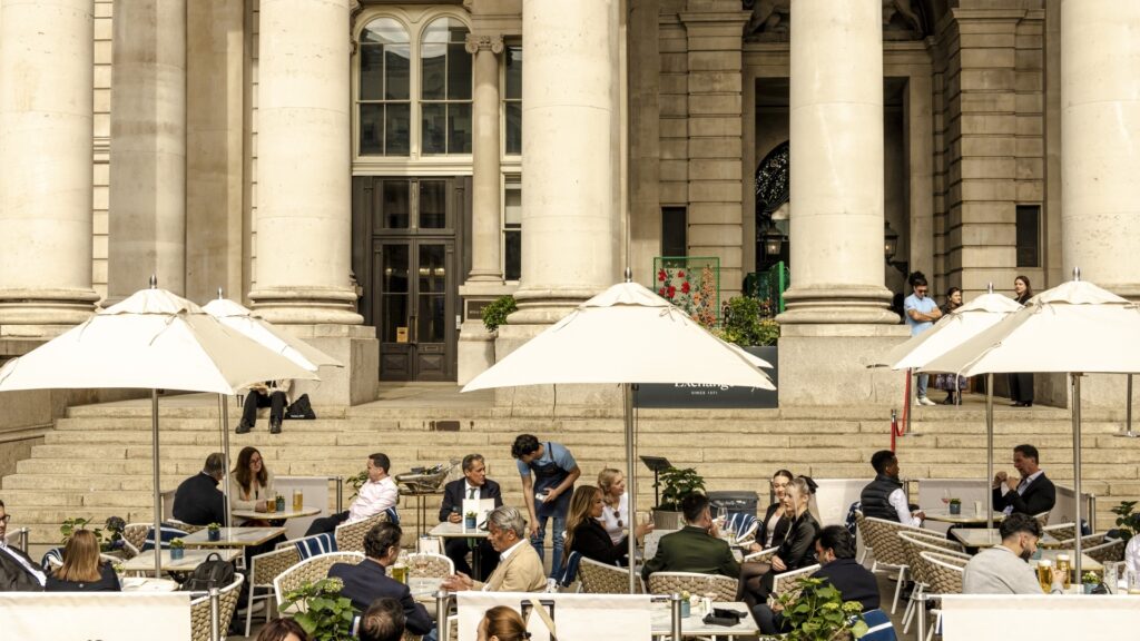 An outdoor terrace with people sitting at tables with umbrellas outside a grand building.