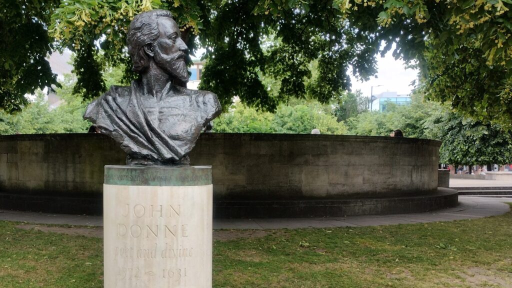 Bust of John Donne on a white stone plinth. Located in a garden with green trees around.