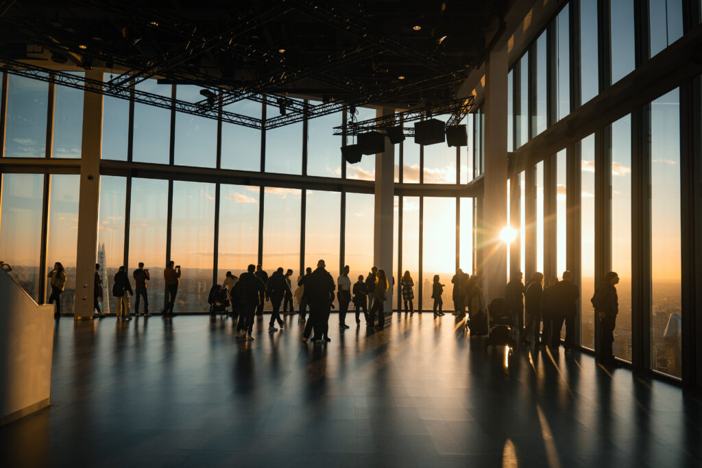 Tall glass windows dominate a spacious area where people stand by the windows, gazing out at the sunset.