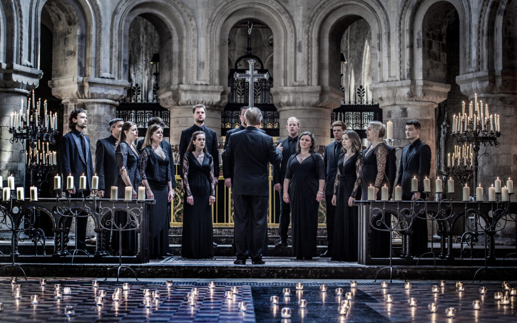 A choir performs in a candle-lit cathedral, directed by a conductor standing in front. The setting features elegant architectural details and reflective flooring, enhancing the atmosphere.