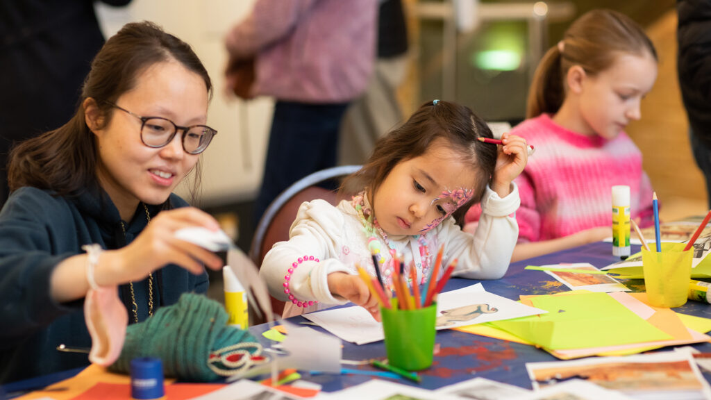 Mother and child engaged in arts and crafts at a table. The mum on the left, wearing glasses, uses a glue bottle. The middle girl with face paint draws, and the girl on the right colors with markers.