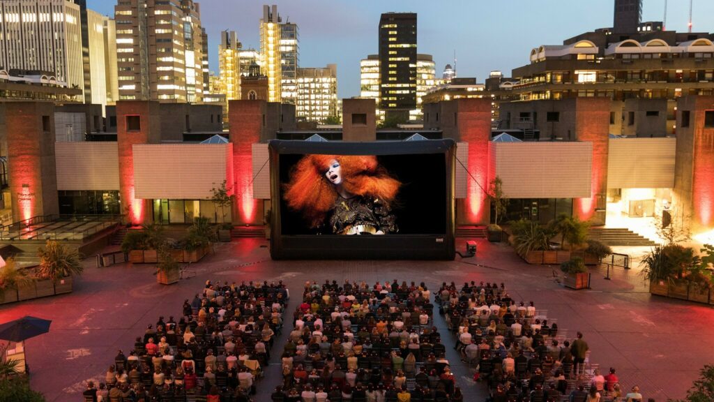 People sitting down on chairs outdoors, watching a giant screen with an image of Bjork.