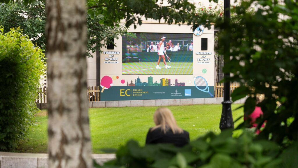 People watching sports on a big screen in a green garden.