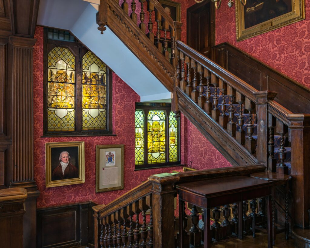 An ornate wooden staircase, stained glass windows, and traditional wall decorations. A chandelier hangs above, and portraits are displayed along the walls