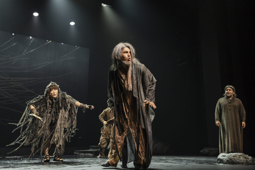 Four actors on stage, dressed in tattered costumes. The set-design is dark with bright spot-lights overhead.