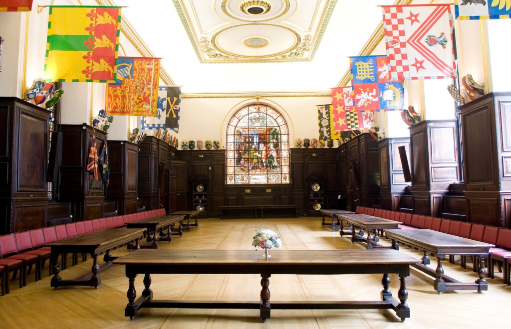 Interior of Stationer’s Hall featuring a long wooden table with a floral centrepiece, surrounded by carved wooden chairs, and decorated with various colourful heraldic banners hanging from the walls. A large stained glass window dominates the far wall under an ornate ceiling.