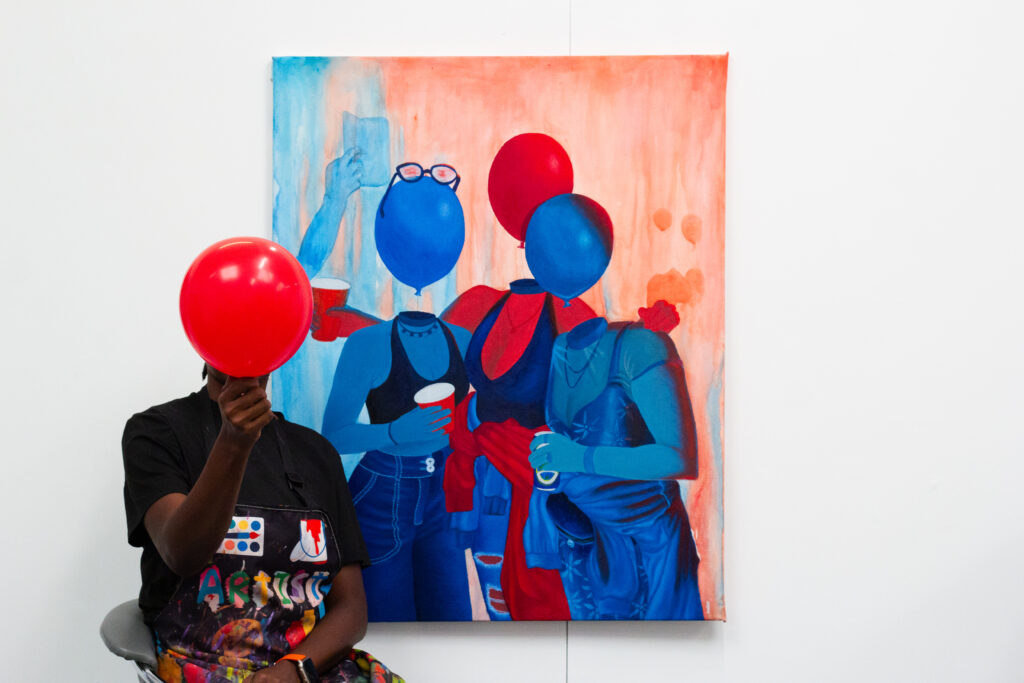 Person holding a red balloon in front of their face, sitting next to a large painting featuring three blue figures with red accents.