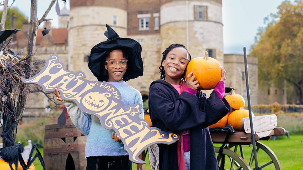 Two children holding a pumpkin and a sign that reads 'Halloween' with a medieval building behind