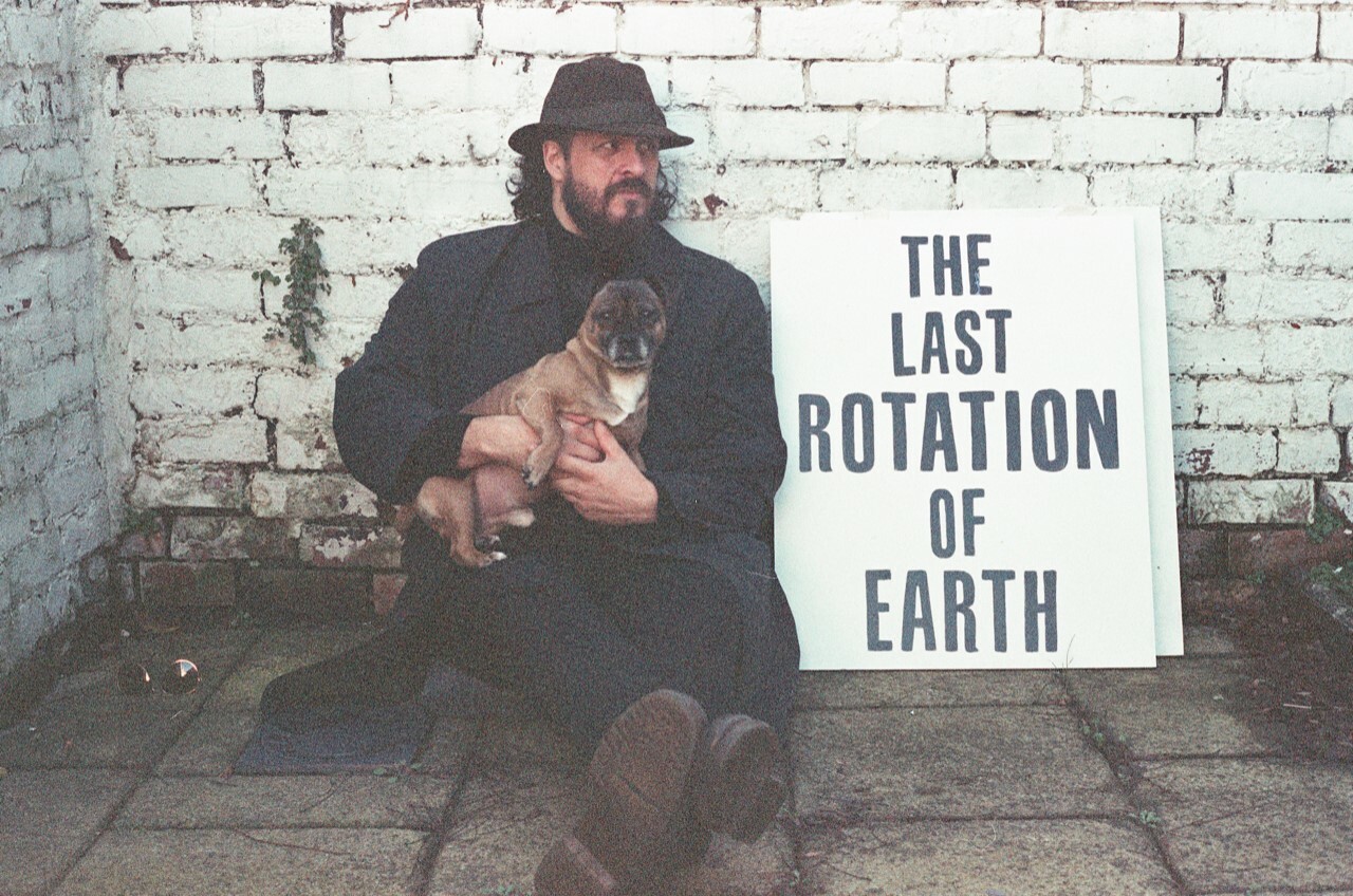 Man sitting on the floor holding a dog, next to a poster with text: The last rotation of earth.