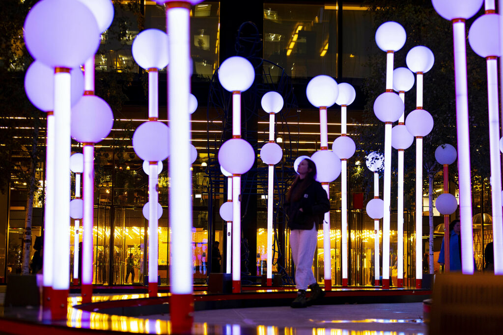 A person stands amidst an outdoor installation with numerous tall, illuminated poles topped with glowing orbs at night, creating a vibrant and futuristic atmosphere.