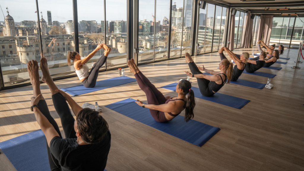 Eight people and a yoga instructor sitting on blue mats in a large room with floor to ceiling windows performing yoga poses. The view outside the window is of the Tower of London.