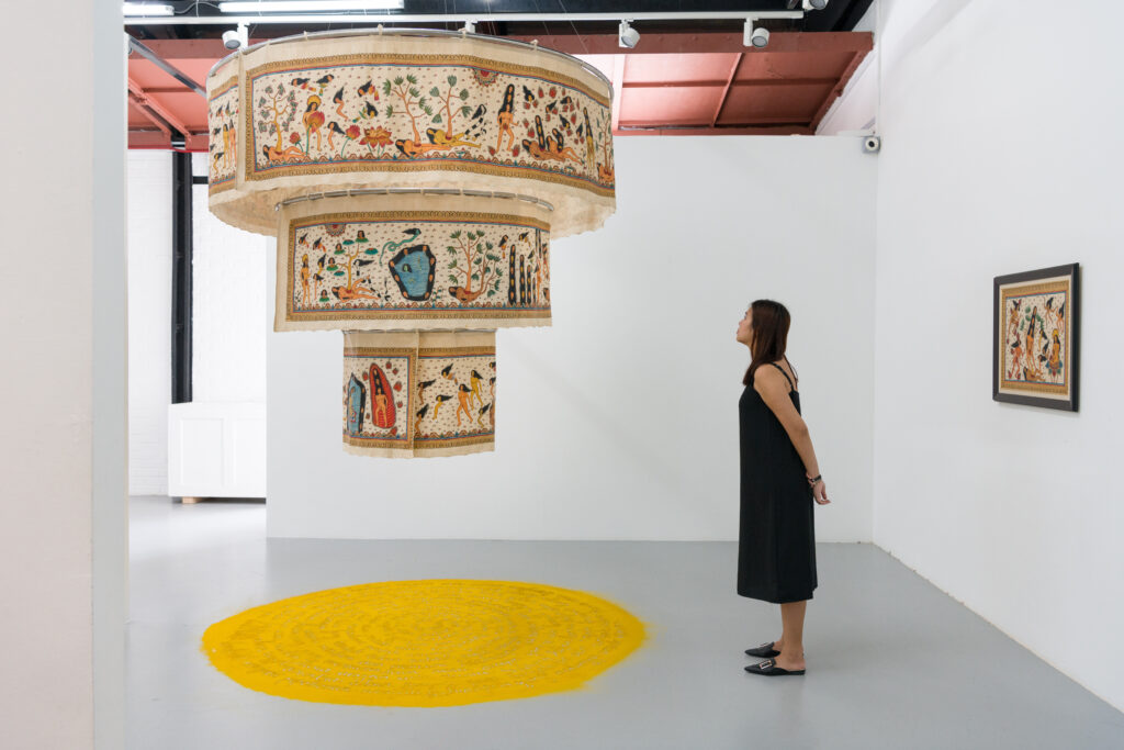 A person stands in an art gallery, observing a suspended, ornate embroidery with a yellow circular rug beneath it. Artworks hang on the white walls around the space.