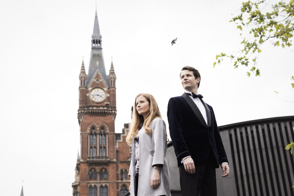 Two individuals in formal attire standing confidently in front of a historical clock tower, with a bird flying in the background.