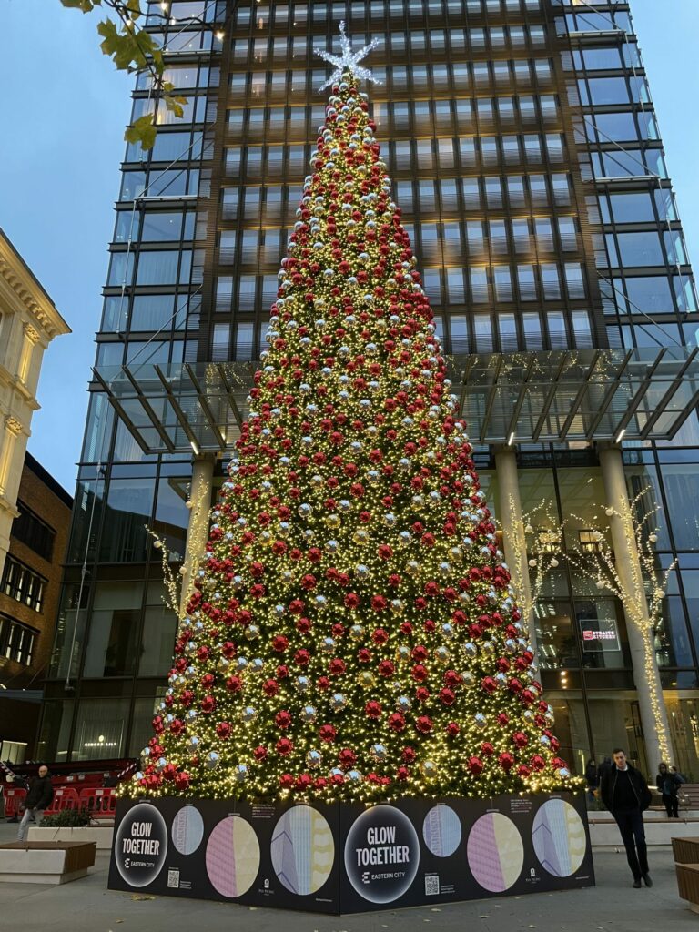 Large Christmas tree outside a modern building, decorated with red and silver baubles, and warm lights.