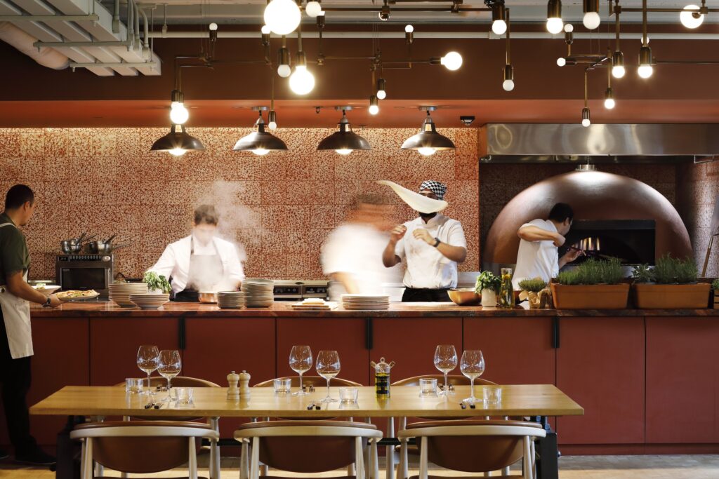 Chefs and staff working in a bustling open kitchen restaurant with a pizza oven, under warm lighting, preparing dishes as diners' tables sit ready in the foreground.