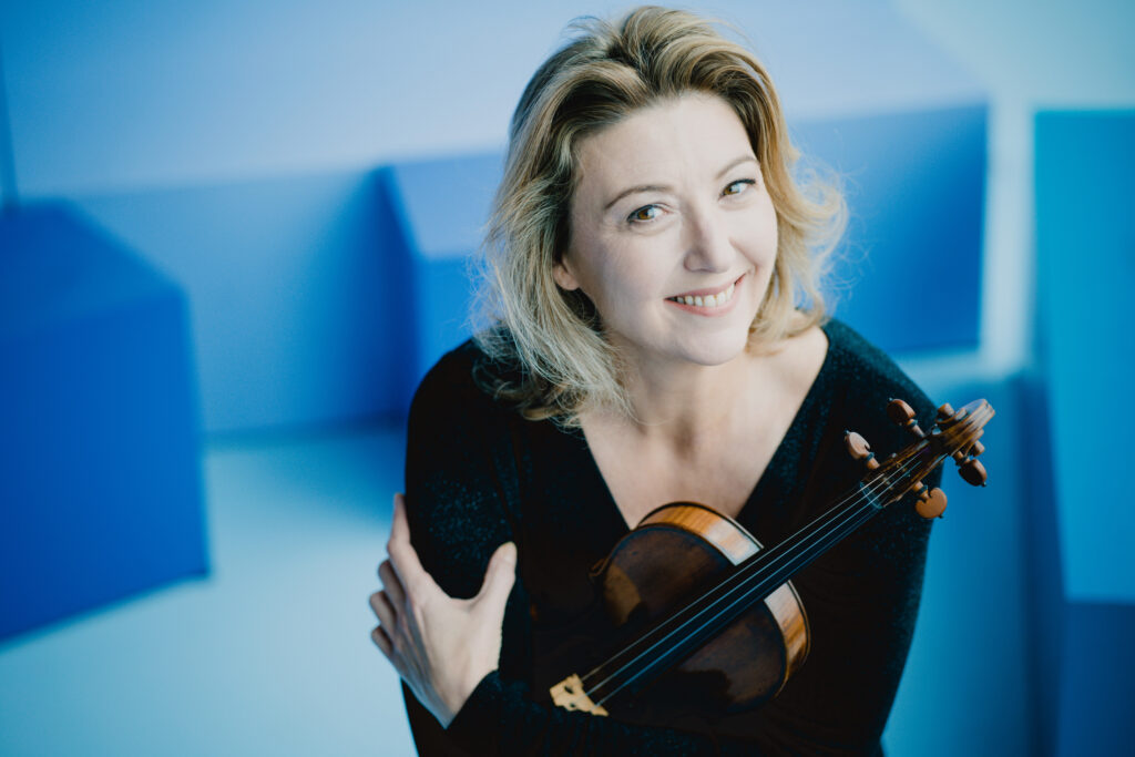 Person holding a violin, smiling in a room with blue geometric shapes in the background.