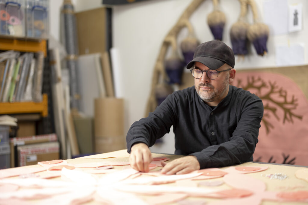 Individual working intently on a large, intricate art project in a studio setting, surrounded by art supplies and a large tapestry in the background.