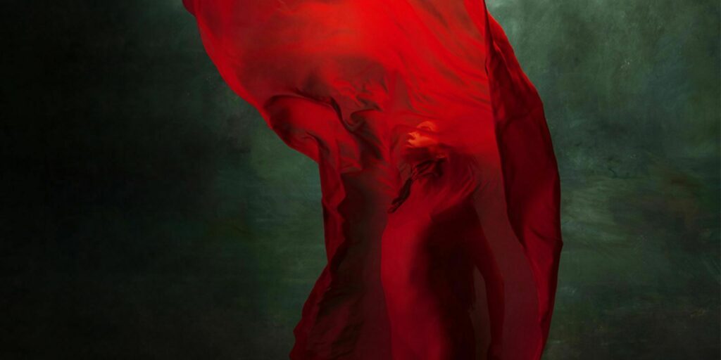 A person enveloped in a flowing red fabric against a dark, textured background.