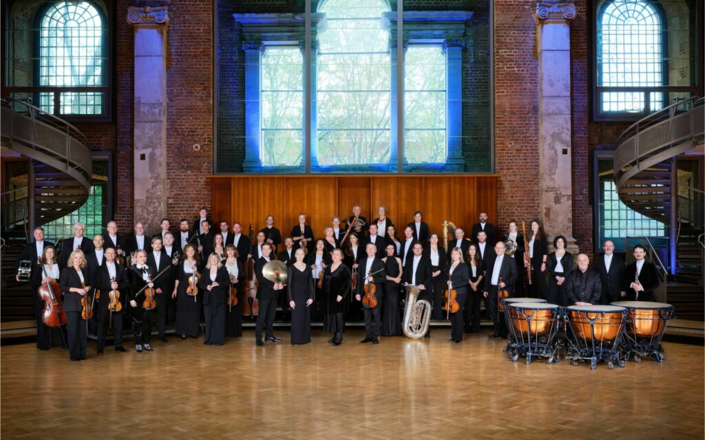 An orchestra posing with their instruments in a grand hall with large windows and brick columns.