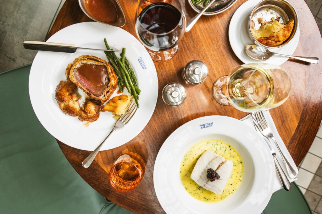 Elegant dining table setup featuring gourmet dishes including roast beef with Yorkshire pudding, asparagus, a fish dish with sauce, and accompanying glasses of red and white wine.