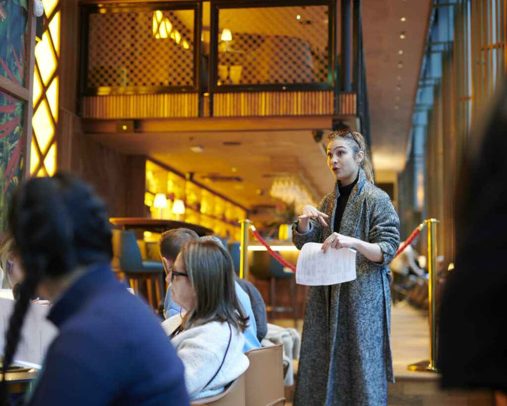 A person in a grey coat conducts an event in a stylish indoor space with wooden furnishings and ambient lighting.