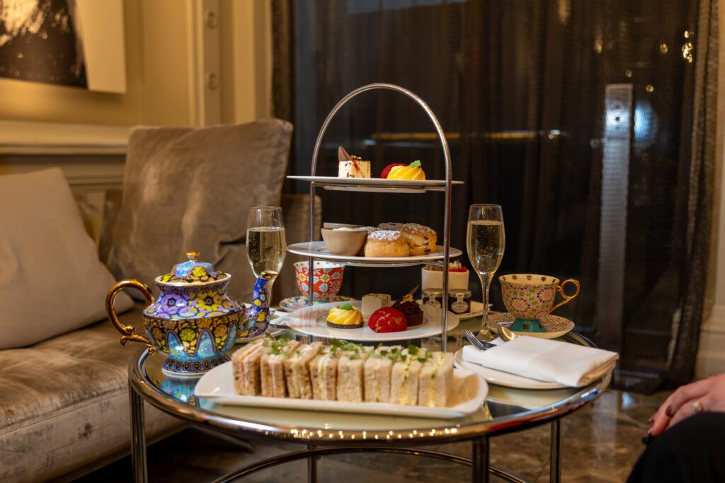 An elegant afternoon tea setup on a glass table, featuring a tiered serving tray with sandwiches, scones, and pastries, complemented by ornate teacups and a glass of champagne.