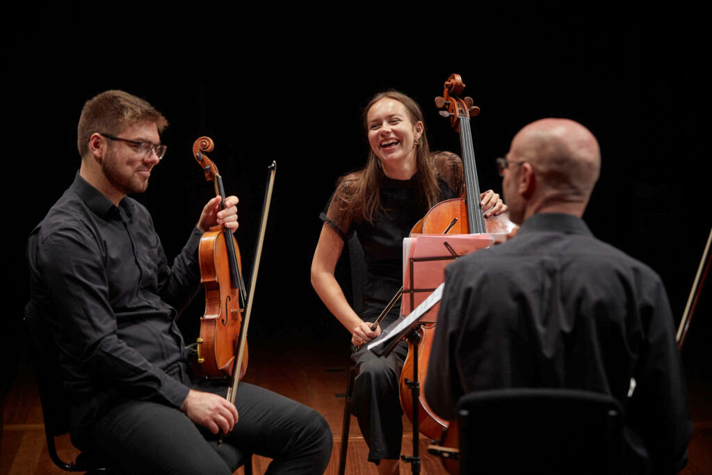 Three cellists, one laughing and two smiling, interact joyfully during a rehearsal on stage.