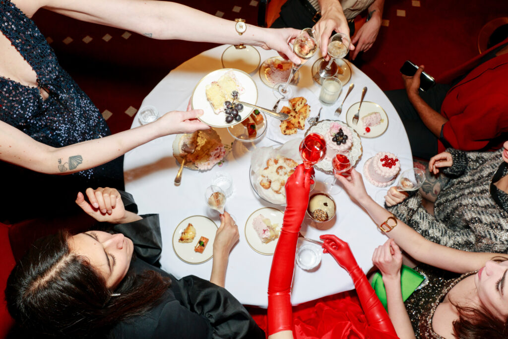 A group of people dining at a table covered with various dishes, reaching towards the center with one person raising a red glass.